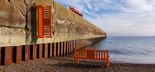 An orange bench on the beach facing the sea with a similar bench vertically attached to a wall