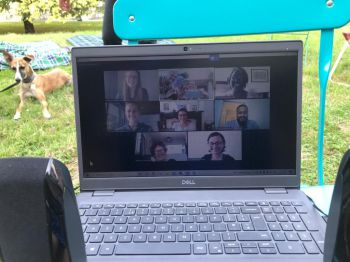 A laptop is shown among physical attendees showing those attending online. A dog lies on the grass behind laptop.