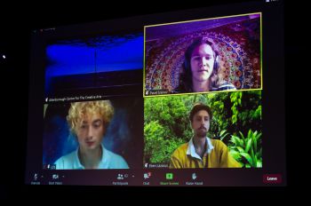 A large screen is divided by four to show three members of a team presenting their idea