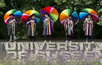 Five students stand atop the concrete University of Sussex sign wearing special rainbow gowns and holding rainbow umbrellas