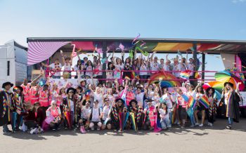 A large group of colourful participants stand in front of a float for the Pride parade