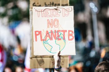 A hand holds up a protest sign reading 'No Planet B' in red hand-drawn lettering, with a drawing of the earth on fire underneath.