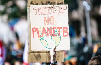 A hand holds up a protest sign reading 'No Planet B' in red hand-drawn lettering, with a drawing of the earth on fire underneath.