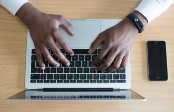 A view from above of a laptop and a mobile phone with two hands reaching out to touch the laptop keyboard