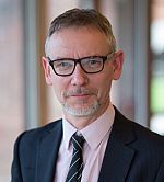Head and shoulders image of Simon Thompson who is facing the camera, wearing a dark suit, pink shirt, dark tie and glasses