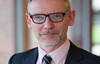 Head and shoulders image of Simon Thompson who is facing the camera, wearing a dark suit, pink shirt, dark tie and glasses