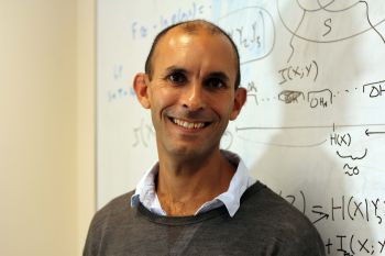 Anil Seth stands before a white board with black writing wearing a grey jumper and light blue shirt