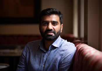 Naimat Zafary pictured sitting on a red couch with his arm resting on its high back wearing a blue shirt