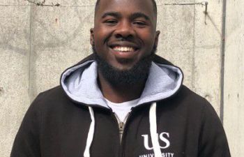 Student Ambassador Shaq Jarrett standing outside a University building in a grey Sussex hoodie.