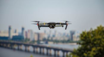 A small drone hovers in the air with a cityscape in the background including a bridge over a wide river