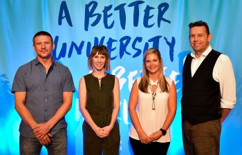 From left to right: Mika Peck, Lucy Hughes,  Melissa Lazenby and Pete Newell stand in front of a light blue backdrop which reads 'A Better University for a Better World'