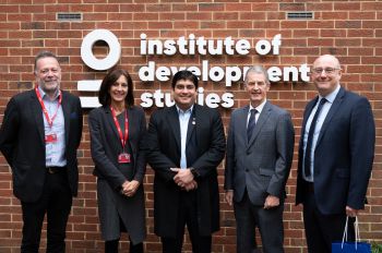 Four men in suits and a woman in a grey dress stand in front of a brick wall with the words Institute of Development Studies