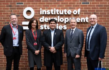 Four men in suits and a woman in a grey dress stand in front of a brick wall with the words Institute of Development Studies
