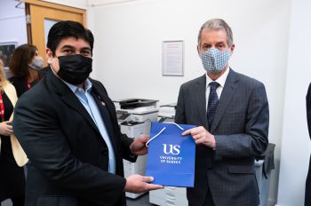 Two men wearing suits exchange a gift while wearing face masks