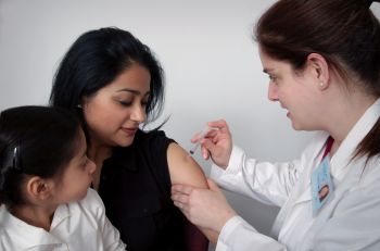 A mother in a black top receives an injection from a doctor while a child in a white shirt sits in her lap