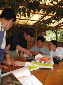 Dr Mika Peck showing four people equipment on a table, as he trains them as parabiologists. Above them is tin roofing with vines draping down.