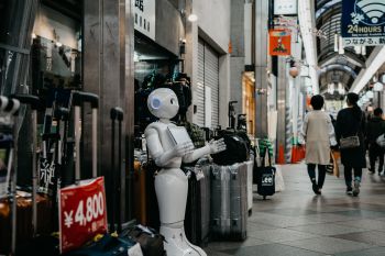 A white robot stands outside a modest shop in a precinct as human shoppers pass by