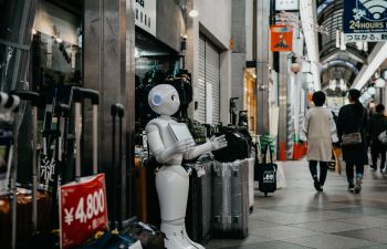 A white robot stands outside a modest shop in a precinct as human shoppers pass by