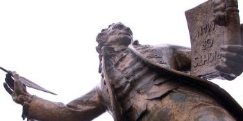 A cropped view of a bronze statue as seen from below. The male figure is dressed in an 18th Century frock coat, waistcoat and flowing neck tie. He holds a feather quill outstretched whilst gripping a book titled 'rights of man' in his left hand.