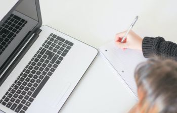 An open laptop on a table next to a woman writing in a notebook