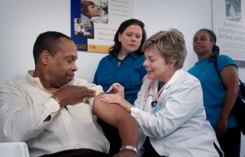 A man of colour rolls up a sleeve to receive a injection from a white female doctor while another healthcare staff member and an apparent family member look on