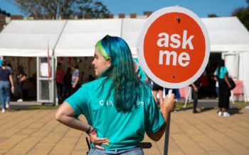 Student ambassador wearing a turquoise t-shirt and matching hair holds an orange round sign that reads