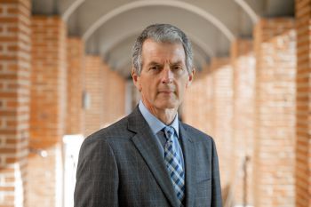 David Maguire stands in a covered walkway with arches staring at the camera wearing a grey suit, shirt and tie