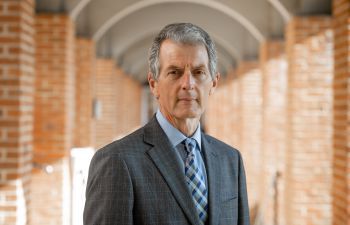 David Maguire stands in a covered walkway with arches staring at the camera wearing a grey suit, shirt and tie