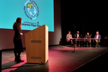 A student stands at a lecturn answering questions from a panel of judges sitting at a table on the other side of the stage