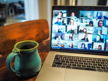 picture of a laptop showing people on a zoom call. a mug sits next to the laptop. both are on a wooden table.