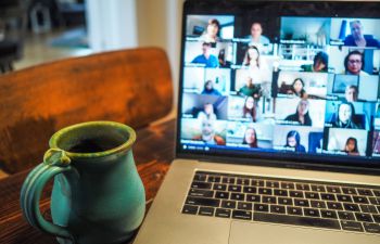 picture of a laptop showing people on a zoom call. a mug sits next to the laptop. both are on a wooden table.