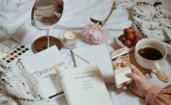 A mobile phone taking a picture of a varied collection of items including strawberries, books about instagram, a mirror, a candle and watercolours.