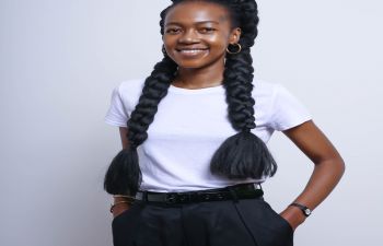 Fumani Mthembi stands with her hands in her pockets wearing a white shirt and black trousers with her hair in two plaits against a plain white backdrop