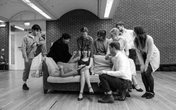 Black and white image of a young woman sitting on a sofa looking at the camera with another girl's head on her lap. A group of people surround the sofa looking at them