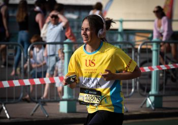 A marathon runner wearing a bright yellow top with University of Sussex logo and giant headphones runs along Brighton seafront