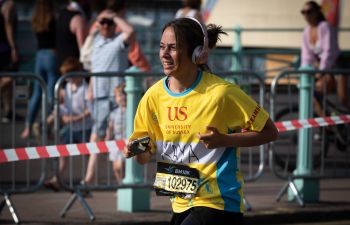 A marathon runner wearing a bright yellow top with University of Sussex logo and giant headphones runs along Brighton seafront