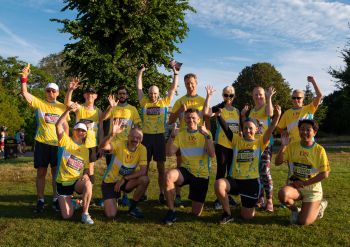 13 runners in bright yellow tops and black shorts, some standing and some kneeling, wave at the camera