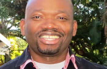 A photo showing Collins Iwuji, a researcher in HIV, smiling at the camera with trees in the background