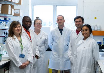 Dr Rhys Morgan (centre) wearing a white lab coat, surrounded by members of his lab, also wearing white coats.