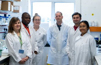 Dr Rhys Morgan (centre) wearing a white lab coat, surrounded by members of his lab, also wearing white coats.
