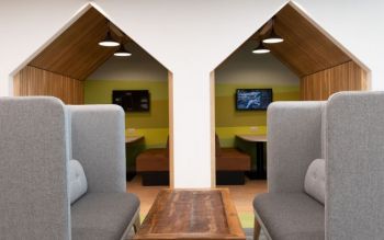 Two grey sofas opposite each other separated by a coffee table. in the background two quiet areas in the shape of beach huts that include sofas, table and display screen