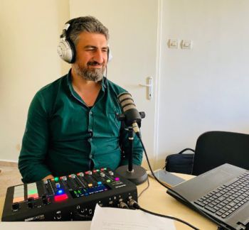 Agricultural Voices Syria podcast host, Eng. Zuhier Agha is pictured sitting at a desk, a microphone in front, wearing headphones and