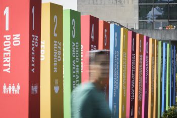 The 17 Sustainable Development Goals presented as separate colourful pillars. A blurry silhouette of a person can be seen walking next to the pillars