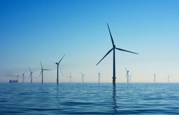 A number of wind turbines rise out of the sea while a boat floats in the background