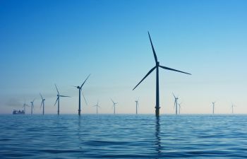 A number of wind turbines rise out of the sea while a boat floats in the background