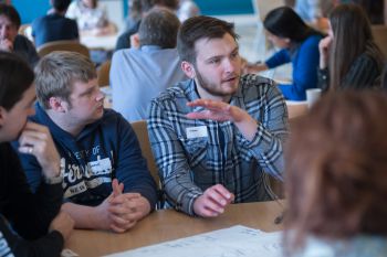 Two attendees sit in their group around a table, with the rest of their group out of shot. One gestures as he adds his opinion to the discussion