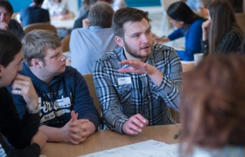 Two attendees sit in their group around a table, with the rest of their group out of shot. One gestures as he adds his opinion to the discussion