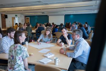 A table of five attendees sit and discuss their project over large sheets of paper at the Biodiversity workshop. In the background you can see 7 more similar tables