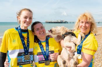Juliet Richardson, Martha Knott and Sam Waugh standing on Brighton beach with their medals. Sam is holding a dog, which looks like it is trying to lick Martha