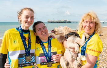 Juliet Richardson, Martha Knott and Sam Waugh standing on Brighton beach with their medals. Sam is holding a dog, which looks like it is trying to lick Martha